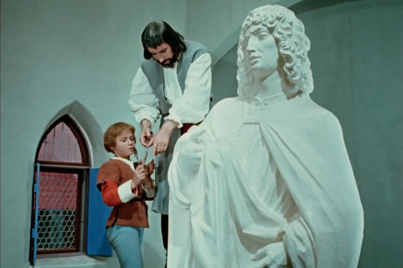 Mężczyzna i dziecko stoją obok dużej, białej statuy. Mężczyzna wydaje się instruować dziecko, trzymając rękę nad ręką dziecka, gdy razem dotykają przedmiotu w pobliżu statuy. W tle widać okno.