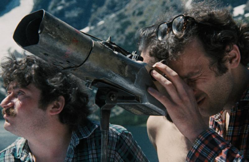 Dwóch mężczyzn na zewnątrz, jeden patrzący przez duży metalowy teleskop, mrużąc oczy, a drugi wpatrzony w dal. W tle widoczne są ośnieżone góry i niebo.