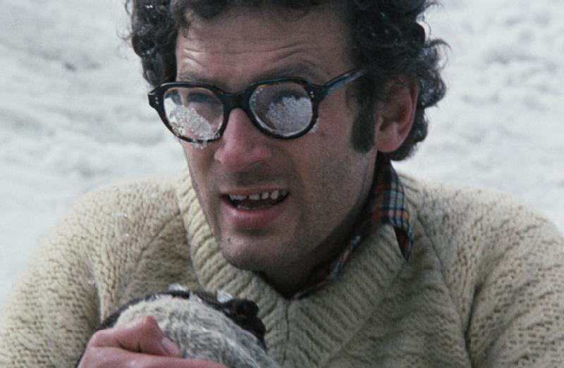 Mężczyzna w grubych okularach z lodem lub śniegiem na soczewkach siedzi na zewnątrz w śniegu, trzymając coś futrzanego. Ma kręcone włosy, kraciastą koszulę i sweter z dzianiny.