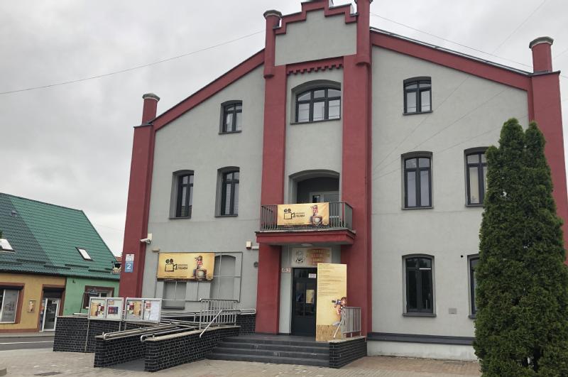 Trzypiętrowy szaro-czerwony budynek z centralnym wejściem, balkonami i podjazdami dla osób niepełnosprawnych. Na balkonie i wejściu znajdują się plakaty i znaki. Obok niego widoczny jest budynek z zielonym dachem i wysokie drzewo.