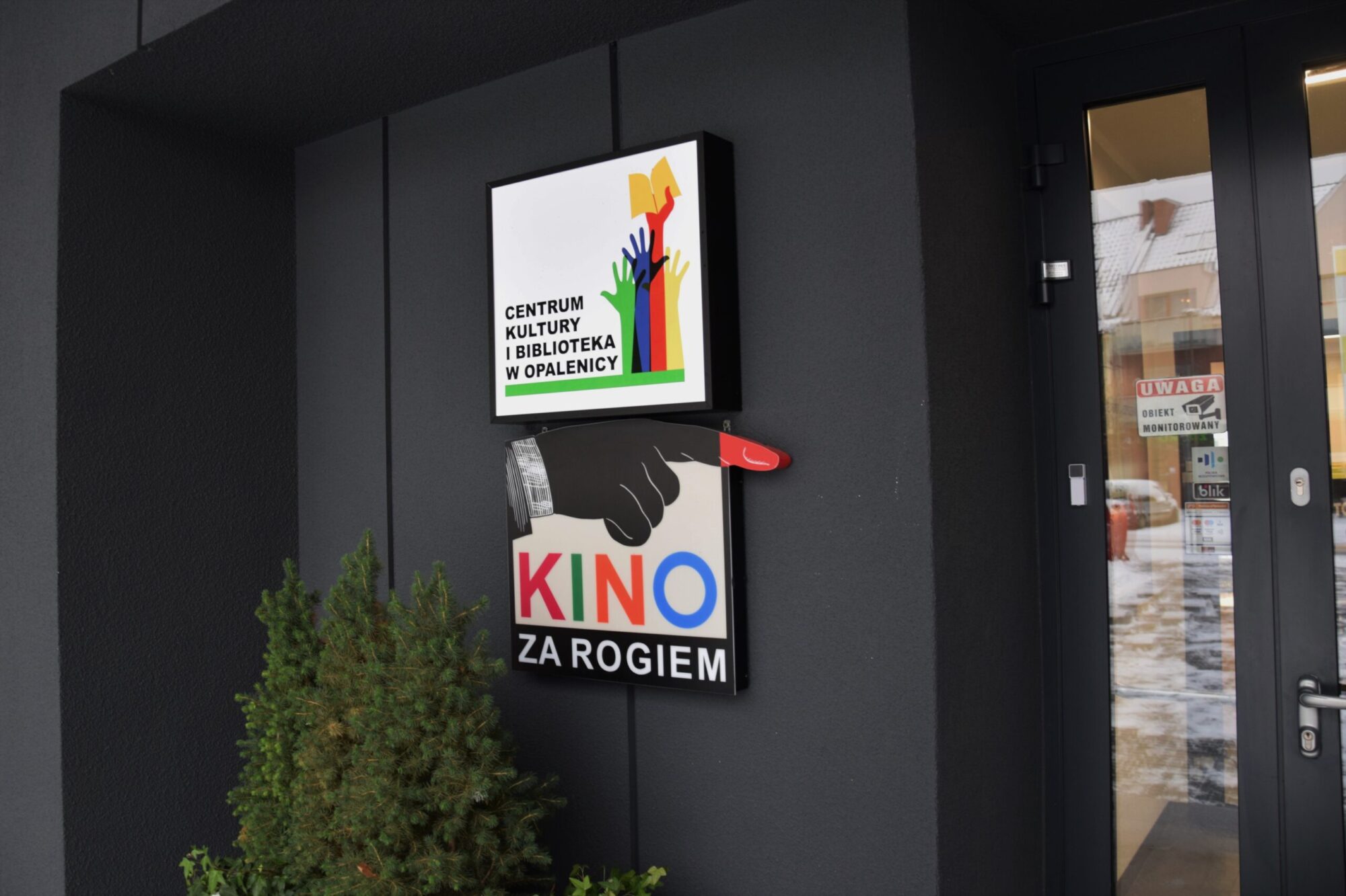 Dwa znaki na ciemnej ścianie w pobliżu drzwi i rośliny. Górny znak przedstawia kolorowe dłonie i głosi Centrum Kultury i Biblioteka w Opalenicy. Dolny znak mówi Kino Za Rogiem z wskazującą dłonią.
