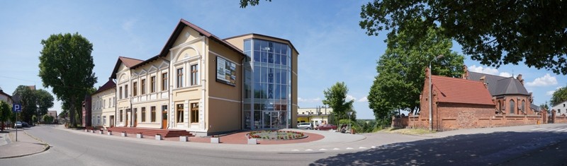 Szeroki widok ulicy ukazuje mieszankę starej i nowoczesnej architektury, na którą składają się zabytkowy beżowy budynek z ozdobnymi detalami obok przeszklonej konstrukcji oraz mały kościół z czerwonej cegły otoczony drzewami pod błękitnym niebem.