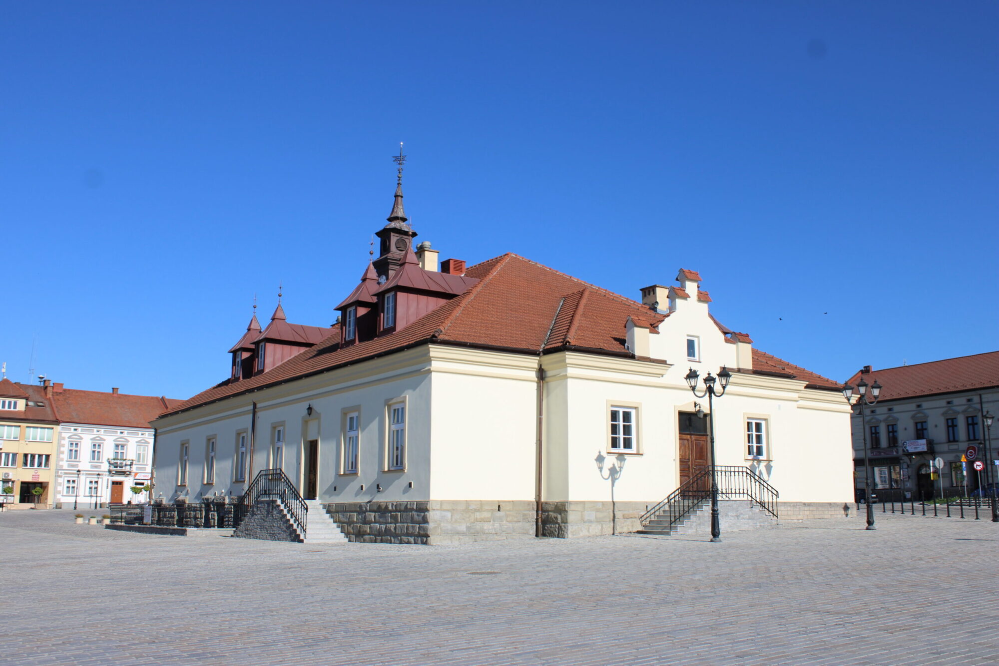 Historyczny, kremowy budynek z dachem z czerwonej dachówki i małymi wieżami stoi na otwartym, brukowanym placu miejskim pod czystym, błękitnym niebem. Schody i zabytkowe latarnie wyznaczają wejście.