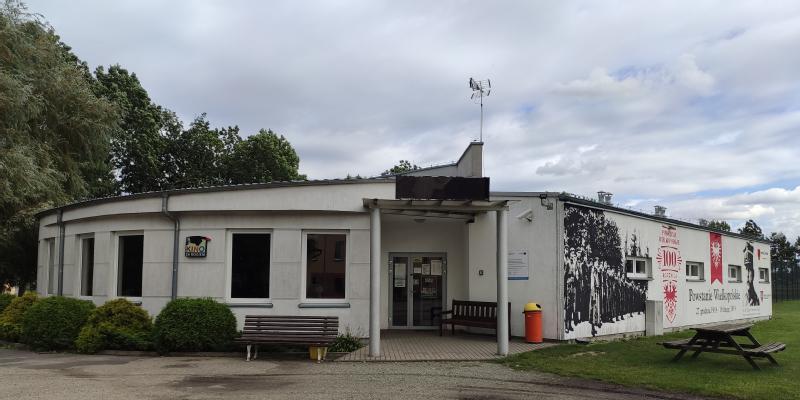 Biały, jednopiętrowy budynek z płaskim dachem i graficznym muralem na jednej ze ścian stoi pośród zieleni. Ławki, kosz na śmieci i zachmurzone niebo są widoczne na zewnątrz wejścia.