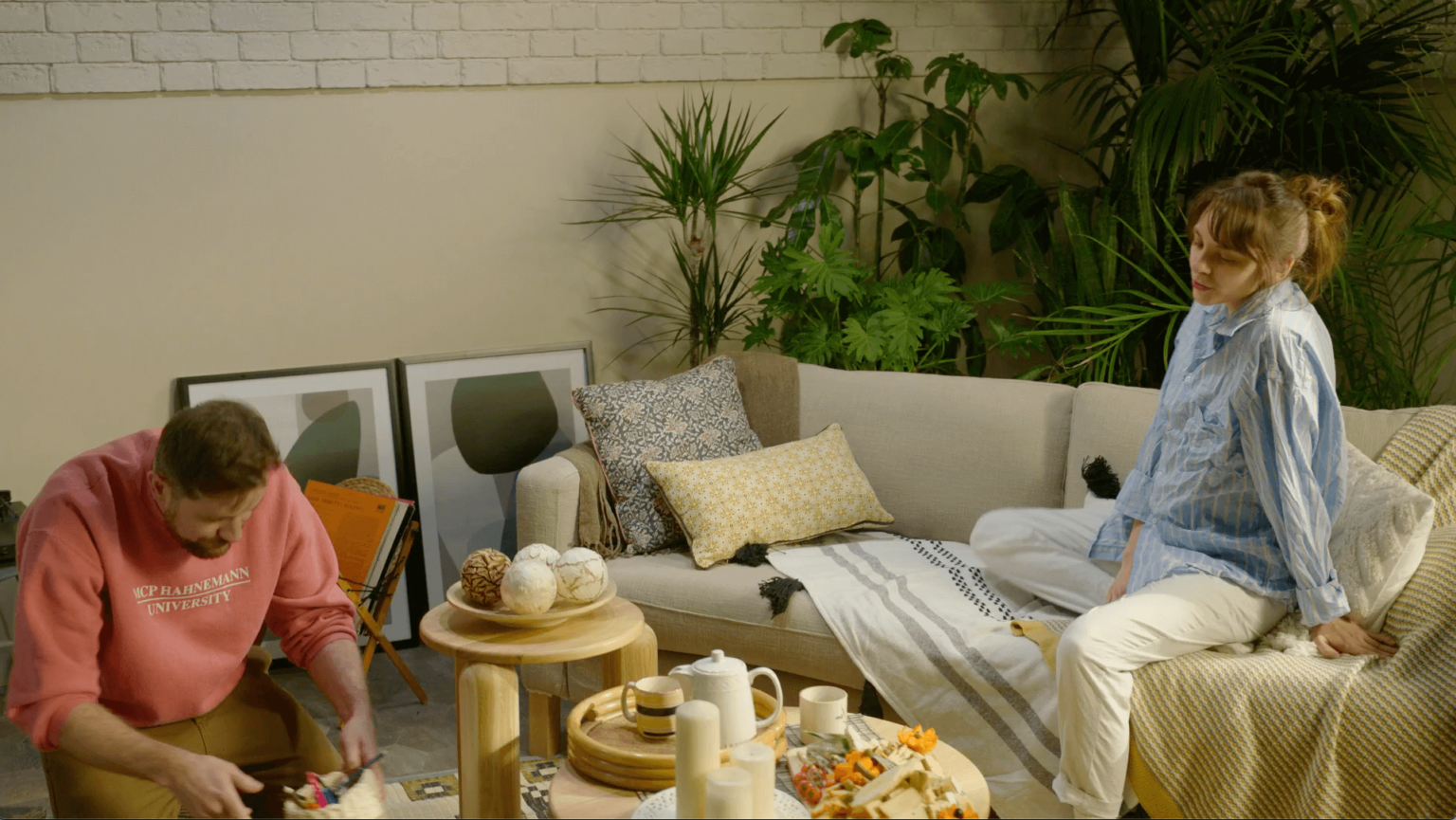 Mężczyzna w różowej bluzie klęczy przy stoliku kawowym i układa przedmioty, podczas gdy kobieta w pasiastej koszuli i białych spodniach siedzi na beżowej sofie. Pokój jest udekorowany roślinami, zdjęciami i przekąskami na stole.