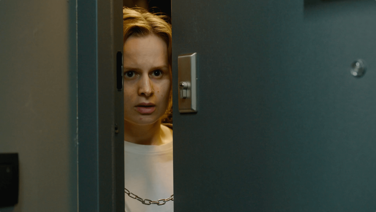 Kobieta o zaniepokojonym wyrazie twarzy patrzy przez częściowo uchylone, zamknięte na łańcuch drzwi, na tle drzwi i ich otoczenia w słabo oświetlonym pomieszczeniu.