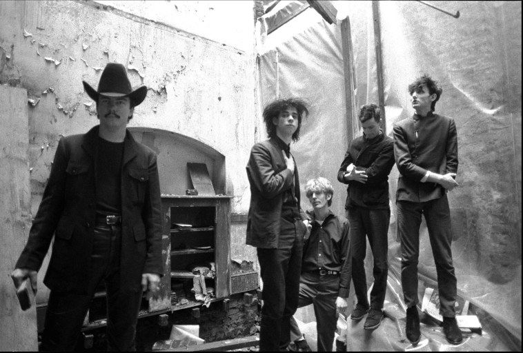 Pięciu mężczyzn ubranych na ciemno stoi w podupadłym pokoju o łuszczących się ścianach; jeden z nich ma na głowie kowbojski kapelusz, a pozostali stoją lub pochylają się swobodnie, niektórzy wyglądają na zamyślonych, w ponurym, miejskim otoczeniu.
