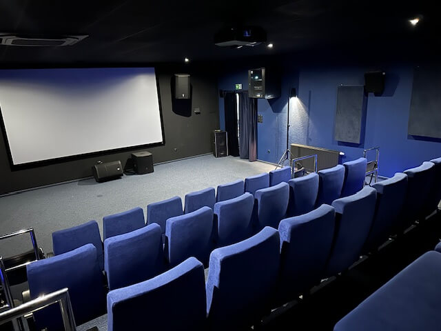 Małe, puste kino z niebieskimi krzesłami zwróconymi w stronę dużego, pustego ekranu projekcyjnego na ciemnoniebieskiej ścianie. Pomieszczenie jest słabo oświetlone, a sprzęt audio jest widoczny w pobliżu ekranu.