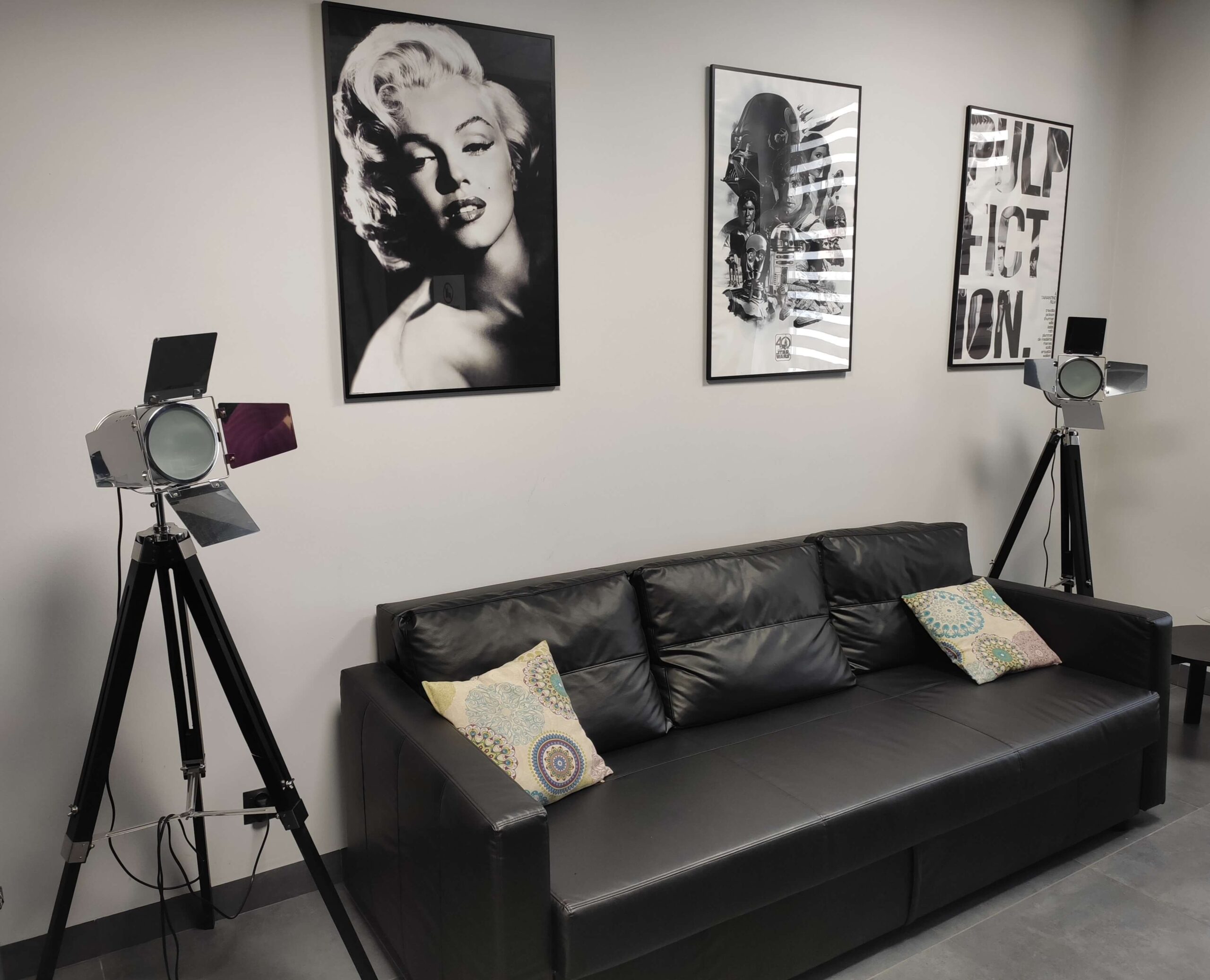 Czarna skórzana kanapa z dwiema ozdobnymi poduszkami stoi przy ścianie, nad którą wiszą trzy czarno-białe, oprawione wydruki artystyczne. Po obu stronach kanapy znajdują się dwa lampy studyjne w stylu vintage na statywach.