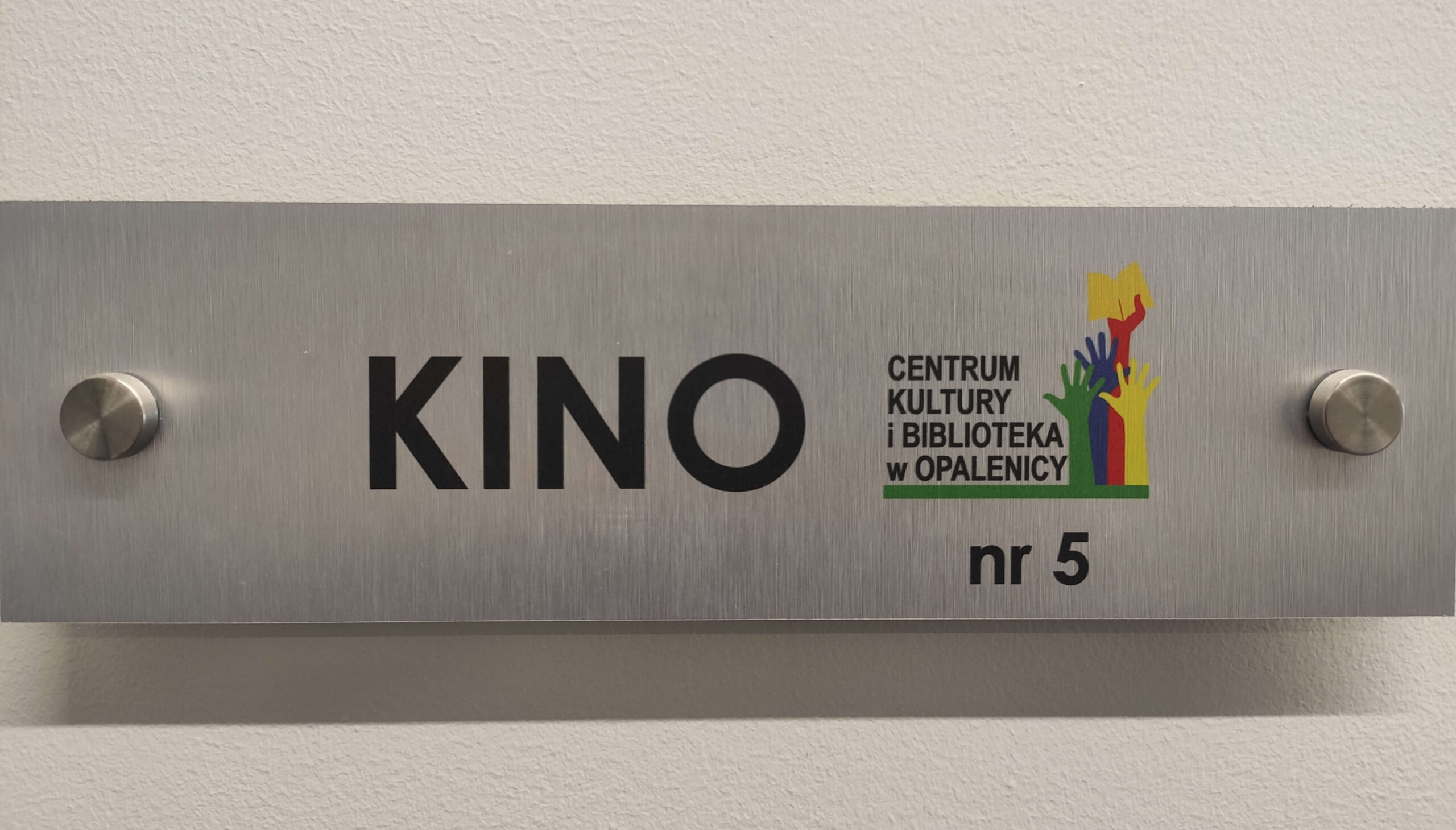 Metalowy szyld na ścianie z napisem KINO nr 5 i Centrum Kultury i Biblioteka w Opalenicy oraz kolorowym logo przedstawiającym uniesione ręce.