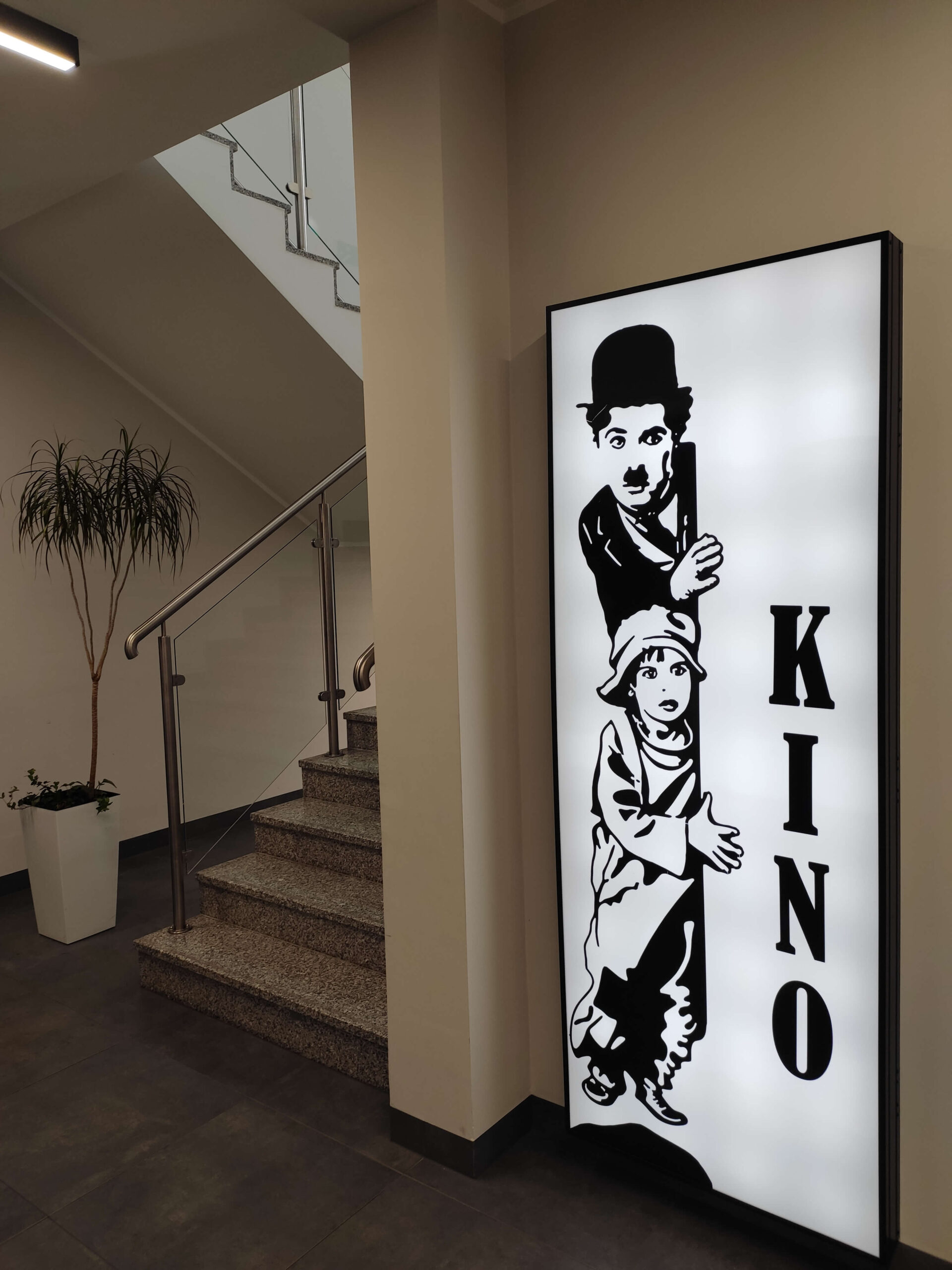 Oświetlony znak przy schodach pokazuje czarno-białe rysunki Charliego Chaplina i dziecka wyglądającego zza słupa. Obok nich pionowo napisane jest słowo KINO. Nieopodal stoi doniczkowa roślina.