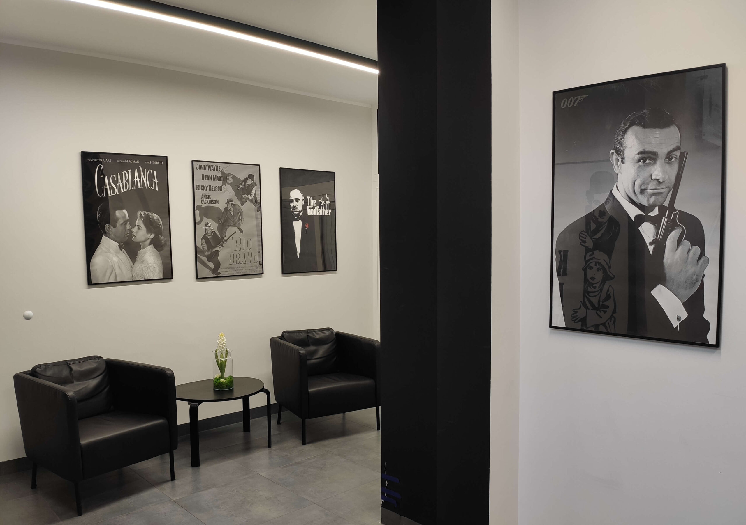 Nowoczesna poczekalnia z dwoma czarnymi fotelami, małym okrągłym stolikiem z białym kwiatem i klasycznymi plakatami filmowymi na ścianie, w tym „Casablancą” i dużym plakatem Jamesa Bonda po prawej stronie.