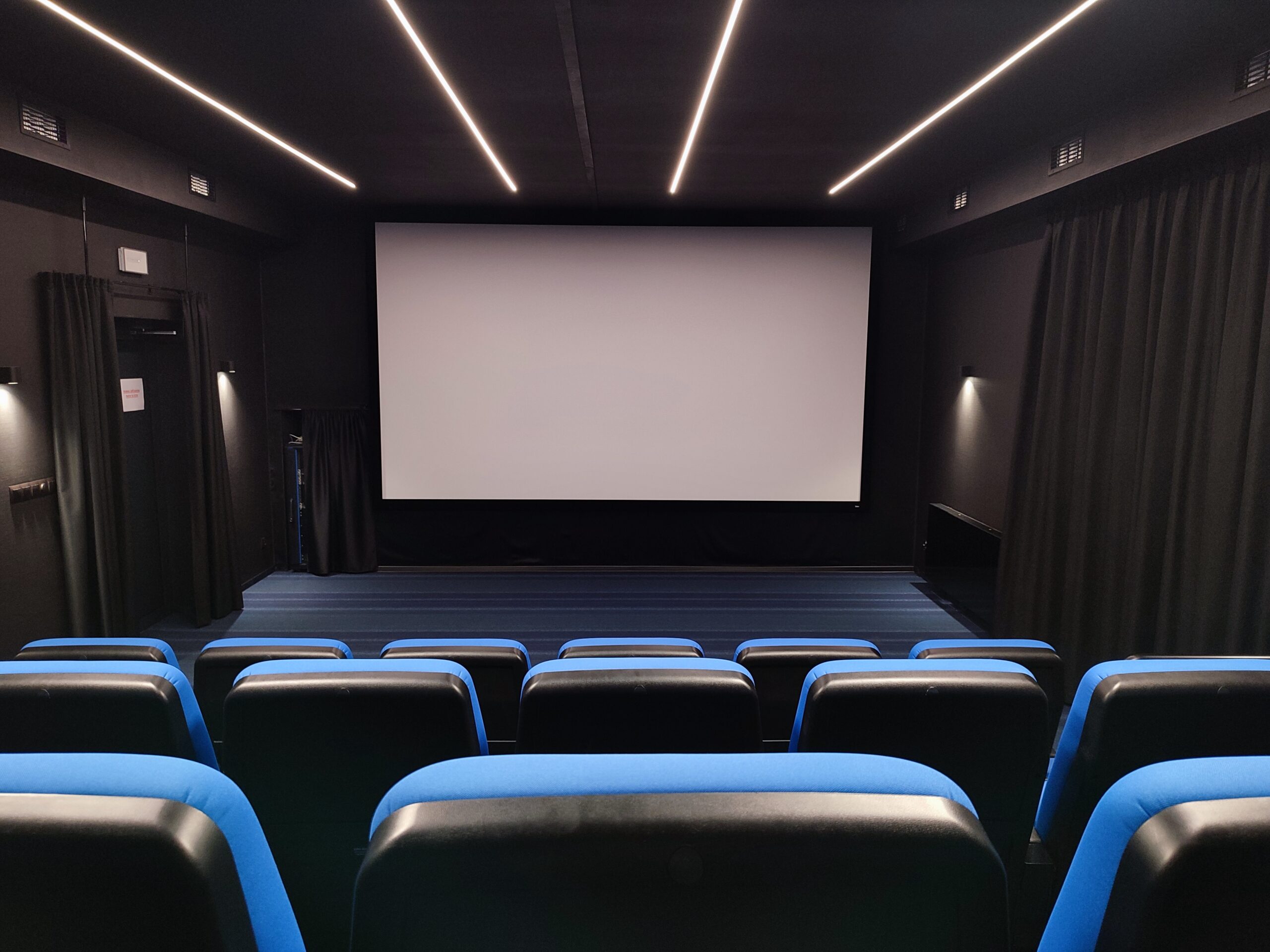 Małe kino z niebieskimi i czarnymi siedzeniami naprzeciwko dużego, pustego białego ekranu. Ciemny pokój ma czarne ściany i sufit, z pionowymi pasami światła nad nimi i zasłonami po bokach.