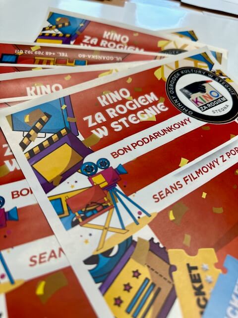 Zbliżenie kolorowych biletów do kina lub voucherów na Kino za Rogiem w Stegnie z ilustracjami kamery filmowej, rolek filmu i ikon prezentów na czerwonym tle. Tekst jest w języku polskim.