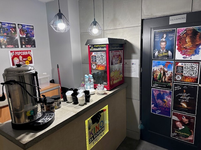 Małe stoisko z przekąskami, maszyną do popcornu, ekspresem do kawy, jednorazowymi kubkami i przyprawami. Na pobliskich ścianach i drzwiach pod nowoczesnymi lampami wiszącymi znajdują się plakaty filmowe i kody QR.
