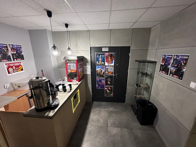 Mały punkt gastronomiczny z maszyną do popcornu, dozownikiem gorących napojów, dwoma wiszącymi lampami, szklaną gablotą i plakatami filmowymi na ścianach i drzwiach. Przestrzeń ma szare ściany i kafelkową podłogę.