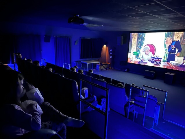 Grupa dzieci siedzi w zaciemnionym kinie, oglądając film animowany na dużym ekranie, który pokazuje postacie z kreskówek przy biurku w jasno oświetlonym pokoju. Niebieskie światło delikatnie oświetla publiczność i pokój.