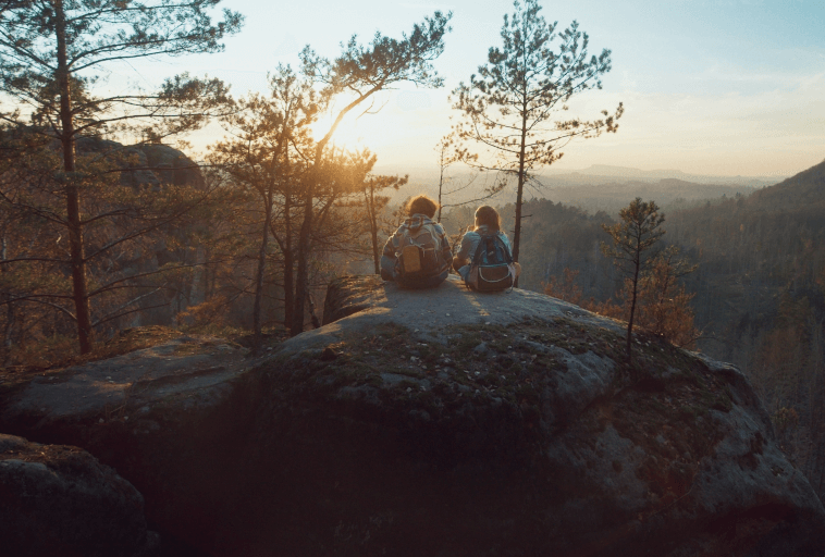 Dwie osoby z plecakami siedzą na dużej skale z widokiem na zalesioną dolinę o zachodzie słońca. Otaczają ich wysokie sosny, a światło słoneczne tworzy ciepłą, spokojną atmosferę.
