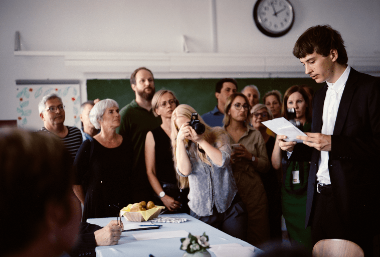 Mężczyzna w garniturze czyta z kartki przed grupą ludzi w klasie. Kobieta fotografuje go, a inni przyglądają się uważnie. W tle widoczny jest zegar i tablica.