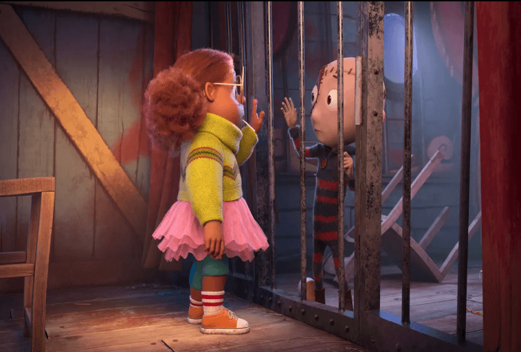 Dziewczyna z kręconymi włosami, w okularach i kolorowym stroju stoi przy klatce, dotykając rąk przez kraty z chłopcem w środku. Scena rozgrywa się w słabo oświetlonym, drewnianym pokoju z belkami i kotwicą w tle.