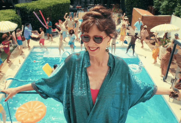 Kobieta w okularach przeciwsłonecznych i błyszczącym niebieskim okryciu uśmiecha się przy basenie, a w tle tańczą i bawią się ludzie. Scena jest jasna i świąteczna, z pływakami basenowymi i kolorowymi parasolami.