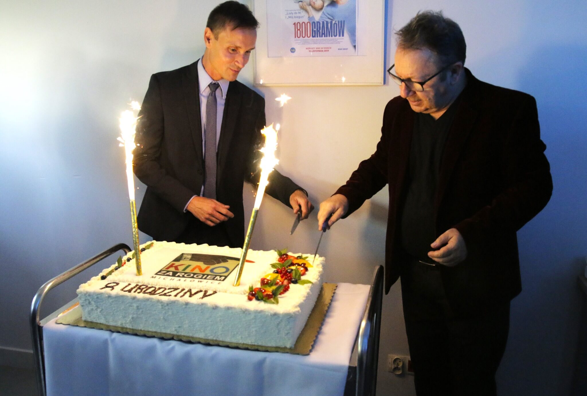 Dwóch mężczyzn stoi obok ozdobionego tortu z zimnymi ogniami, przygotowując się do jego pokrojenia. Tort zawiera słowa „KINO” i „2 URODZINY”, wskazujące na obchody drugiej rocznicy. Na ścianie za nimi widoczny jest plakat.