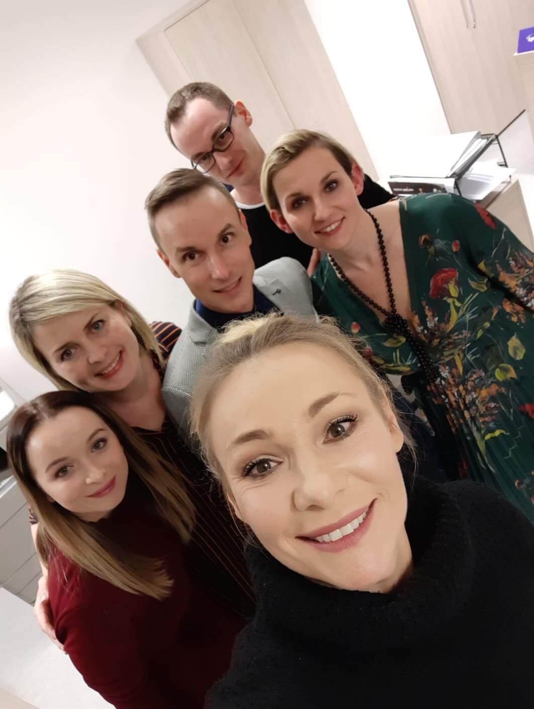 Grupa sześciu dorosłych, trzech mężczyzn i trzy kobiety, uśmiechają się i pozują blisko siebie do selfie w dobrze oświetlonym pomieszczeniu, na tle jasnych ścian i mebli.