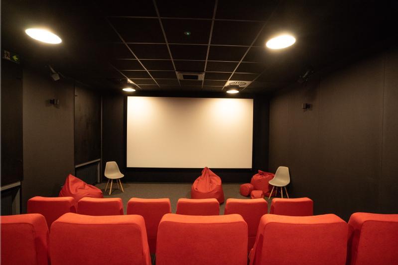 Niewielkie, nowoczesne kino z czerwonymi siedzeniami na pierwszym planie, kilkoma czerwonymi workami wypełnionymi fasolą i białymi krzesłami w pobliżu wejścia oraz dużym, pustym ekranem na ścianie pod oświetleniem sufitowym.