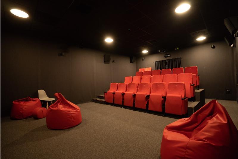 Niewielka sala kinowa z czerwonymi fotelami ustawionymi w rzędach, czerwonymi workami wypełnionymi fasolą i jednym białym krzesłem z przodu, wszystko otoczone ciemnymi ścianami i oświetlone lampami sufitowymi.