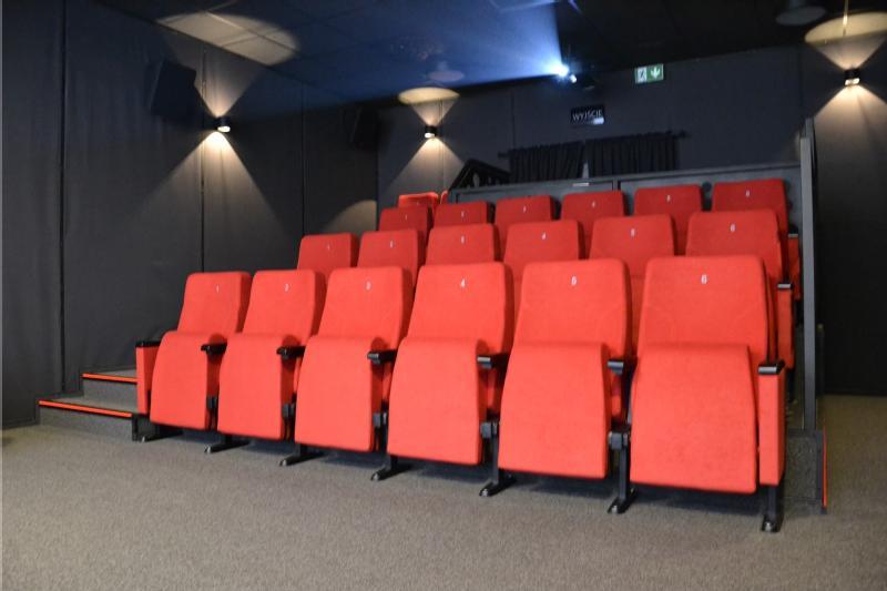 Rzędy pustych czerwonych foteli teatralnych są ustawione w małym, nowoczesnym kinie z ciemnymi ścianami i wykładziną podłogową. Fotele są ponumerowane, a pomieszczenie jest delikatnie oświetlone.