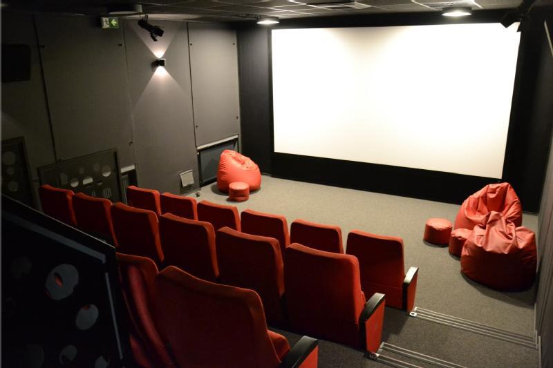Małe, nowoczesne kino z czerwonymi siedzeniami ustawionymi w rzędach, czerwonymi workami fasoli z przodu i dużym pustym ekranem na ścianie. Pomieszczenie jest słabo oświetlone czarnymi i szarymi ścianami.