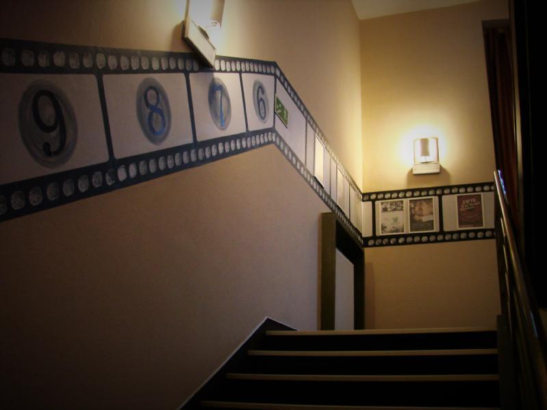 Słabo oświetlone schody z dekoracją ścienną przedstawiającą taśmy filmowe, dużymi liczbami i oprawionymi zdjęciami, prowadzące w górę obok dwóch lamp zamontowanych na ścianach.