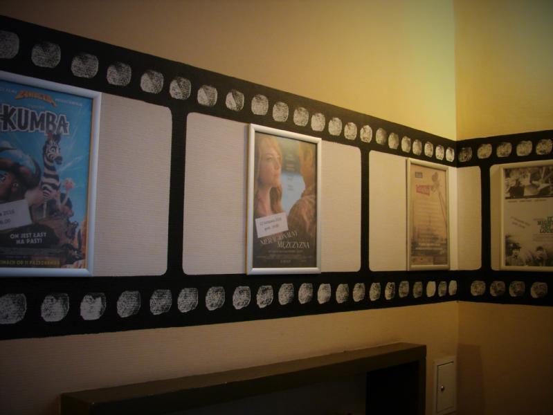 Ściana ozdobiona wzorem taśmy filmowej wyświetla kilka plakatów filmowych, w tym film animowany po lewej i romantyczny dramat pośrodku. Obszar jest ciepło oświetlony z częściowym widokiem na przełącznik światła poniżej.