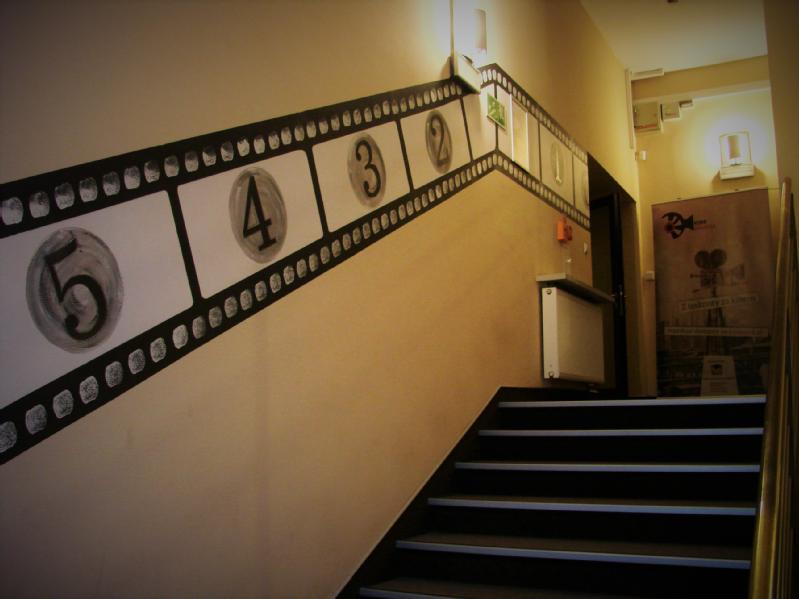 Klatka schodowa z filmową grafiką na ścianie, wyświetlającą odliczanie od 5 do 1. Ściany są beżowe, a schody wyłożone są ciemnymi dywanami. Wzdłuż ściany zamontowano grzejnik i znaki bezpieczeństwa.