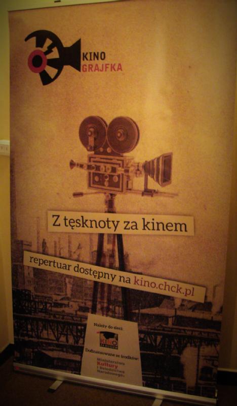 Stojący baner reklamowy kina Grajfka, przedstawiający ilustrację starej kamery filmowej, polski tekst i informacje o stronie internetowej. Tło jest w odcieniach sepii, a dolna część nawiązuje do tematyki przemysłowej.