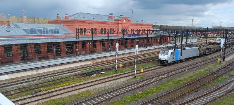 Czerwona ceglana stacja kolejowa z szarym dachem stoi obok wielu torów kolejowych. Nowoczesna biało-niebieska lokomotywa ciągnie wagony towarowe pod zachmurzonym niebem.