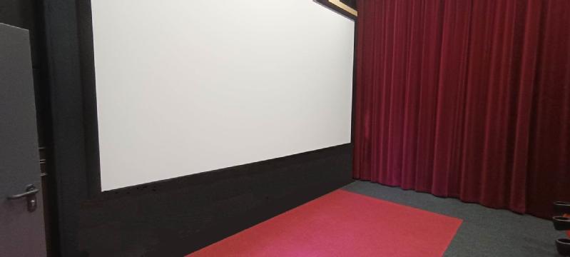 Niewielka sala kinowa z dużym, pustym, białym ekranem, czerwoną wykładziną, czerwonymi zasłonami po prawej stronie i szarymi drzwiami po lewej stronie.