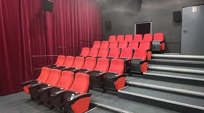 Małe, puste kino z rzędami czerwonych siedzeń, szarą wykładziną, ciemnymi ścianami i bordowymi zasłonami pokrywającymi jedną ścianę. Z tyłu są drzwi i metalowe poręcze wzdłuż przejścia.