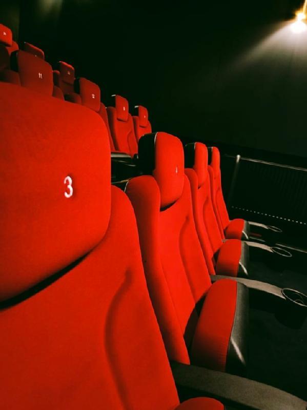 Rzędy pustych czerwonych foteli teatralnych z czarnymi podłokietnikami są pokazane w słabo oświetlonym kinie. Siedzenie najbliżej przodu wyświetla numer 3, a w tle widać więcej ponumerowanych siedzeń.