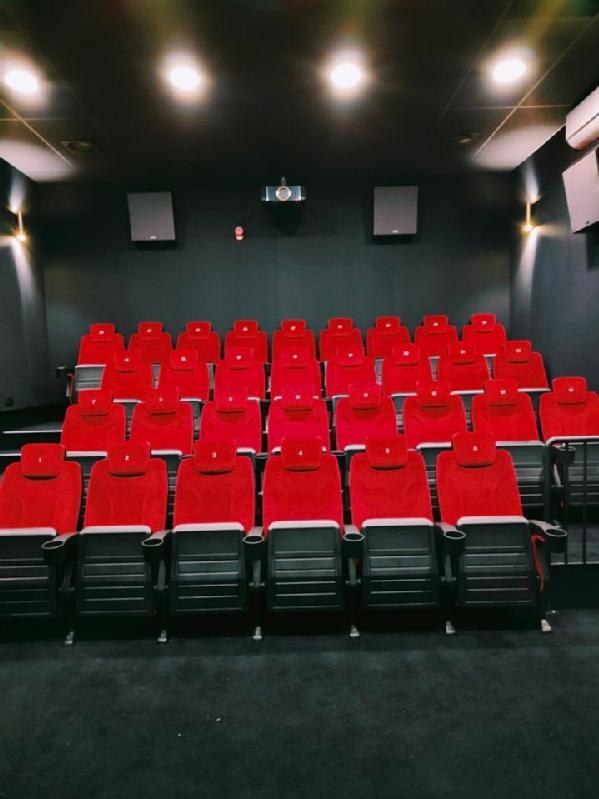 Rzędy pustych, czerwonych siedzeń w teatrze ustawione są naprzeciwko pustego ekranu w słabo oświetlonym, nowoczesnym kinie.
