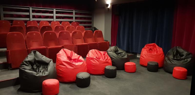 Małe kino z czerwonymi, wyściełanymi siedzeniami w rzędach i kilkoma czerwonymi i czarnymi workami fasolowymi z pasującymi okrągłymi pufami z przodu. W sali jest ciemna wykładzina i niebieskie zasłony przy ekranie.