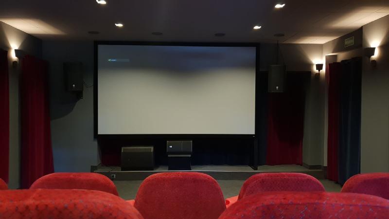 Puste kino z czerwonymi siedzeniami zwróconymi w stronę dużego, pustego ekranu. Włączone są słabe światła ścienne, a po obu stronach ekranu zamontowano głośniki.