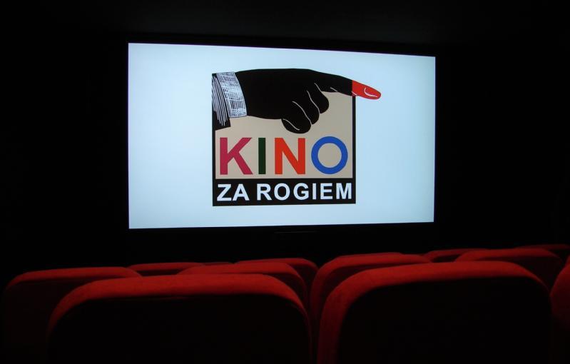 Ekran kina wyświetla logo przedstawiające czarną dłoń wskazującą w prawo z czerwonym czubkiem palca, nad napisem KINO ZA ROGIEM w kolorowych literach. Na pierwszym planie widoczne są czerwone fotele kinowe.