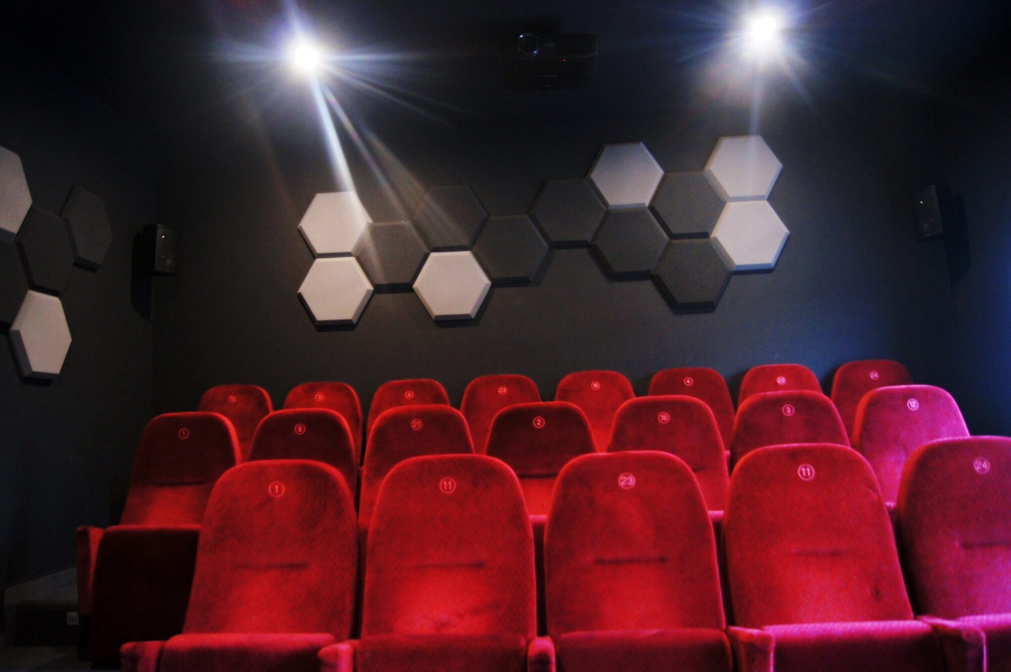 Małe, puste kino z rzędami czerwonych, aksamitnych siedzeń i sześciokątnymi panelami akustycznymi na ciemnej ścianie, oświetlone dwoma jasnymi lampami sufitowymi.