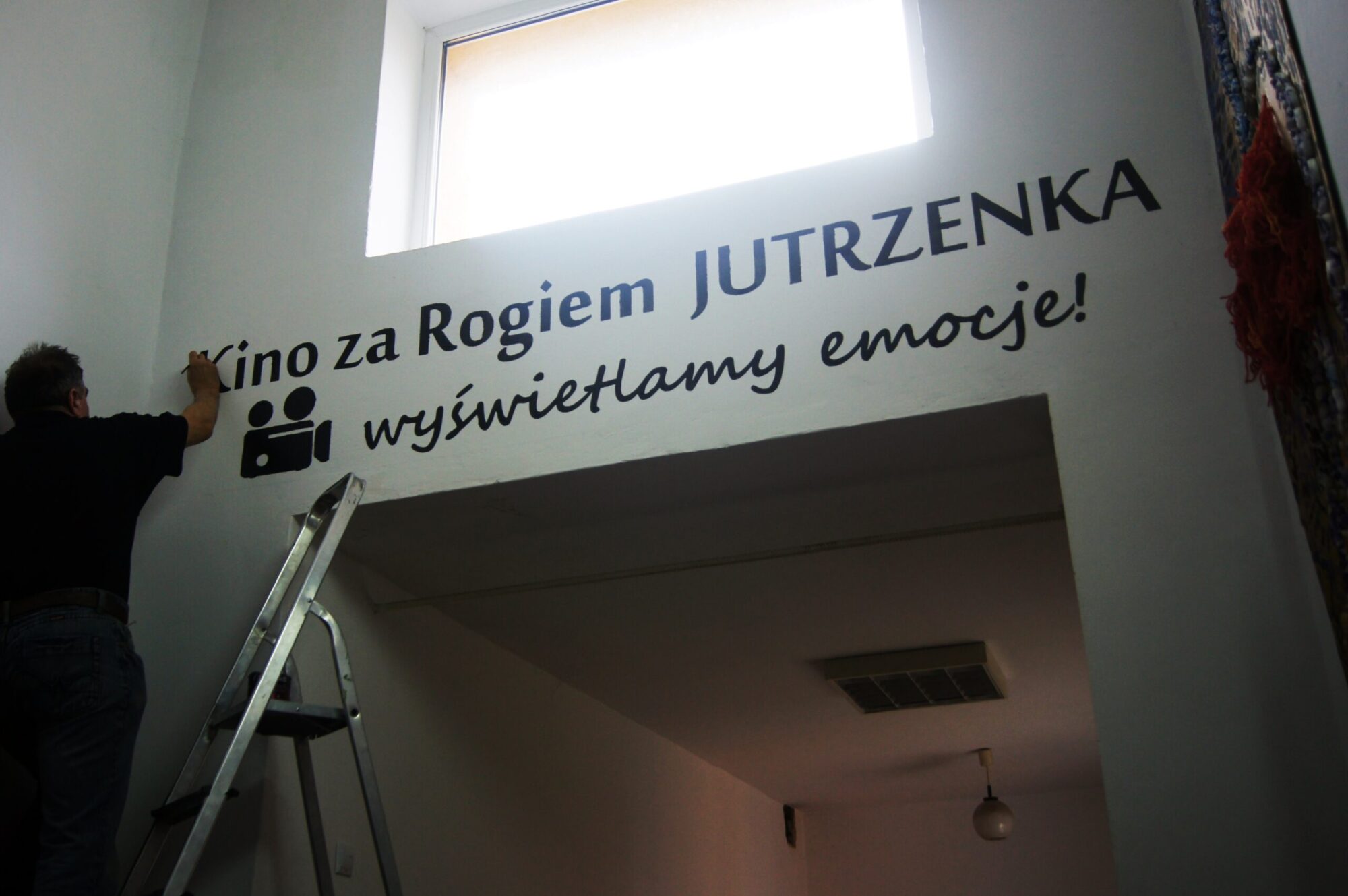 Osoba stoi na drabinie, malując lub instalując polski tekst i ikonę kamery filmowej na białej ścianie pod oknem. Tekst brzmi Kino za Rogiem JUTRZENKA wyświetlamy cierpienie!.