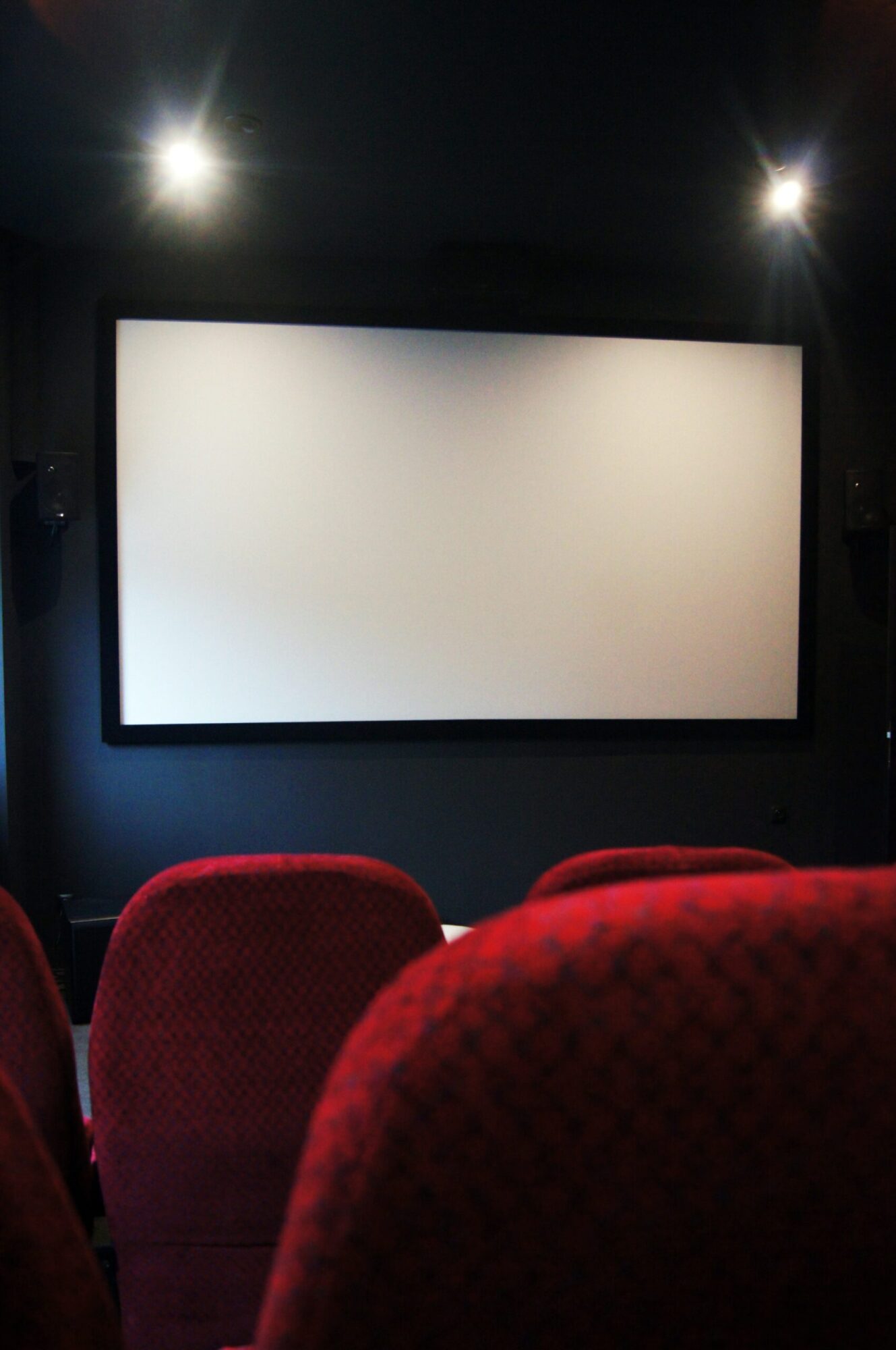 Sala kinowa z pustymi, czerwonymi siedzeniami naprzeciwko dużego, białego ekranu, oświetlonego słabym światłem sufitowym.