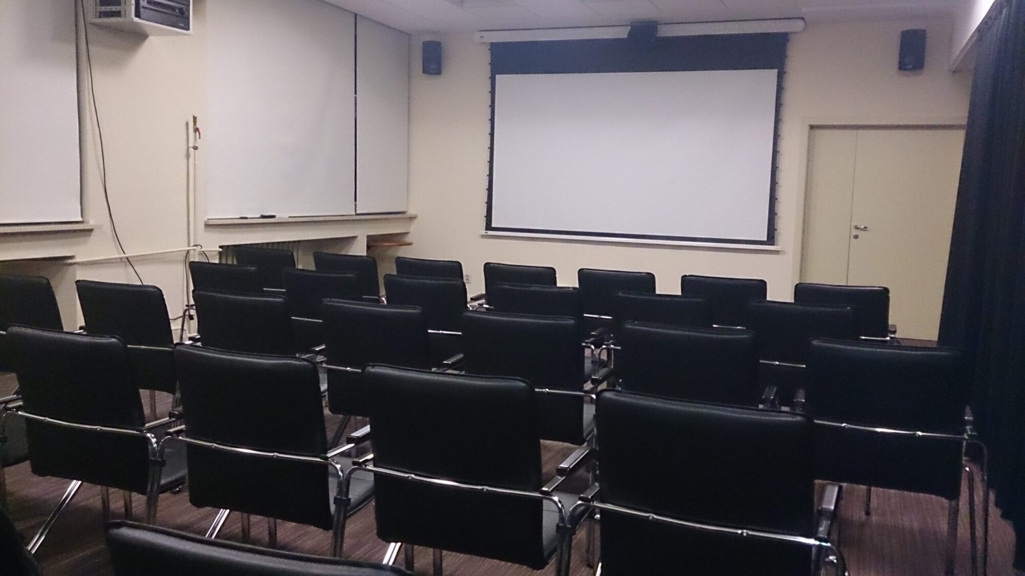 Rzędy pustych czarnych krzeseł ustawione naprzeciwko dużego białego ekranu projekcyjnego w małej, nowoczesnej sali konferencyjnej lub klasowej. W pomieszczeniu znajduje się tablica, beżowe ściany i minimalistyczny wystrój.