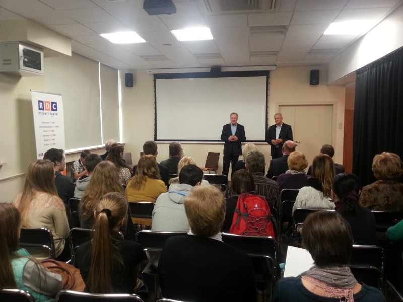 Grupa ludzi siedzi naprzeciwko dwóch mężczyzn stojących z przodu małej sali konferencyjnej. Mężczyźni przemawiają przed pustym ekranem projektora. Po lewej stronie widoczny jest baner z logo RDC.