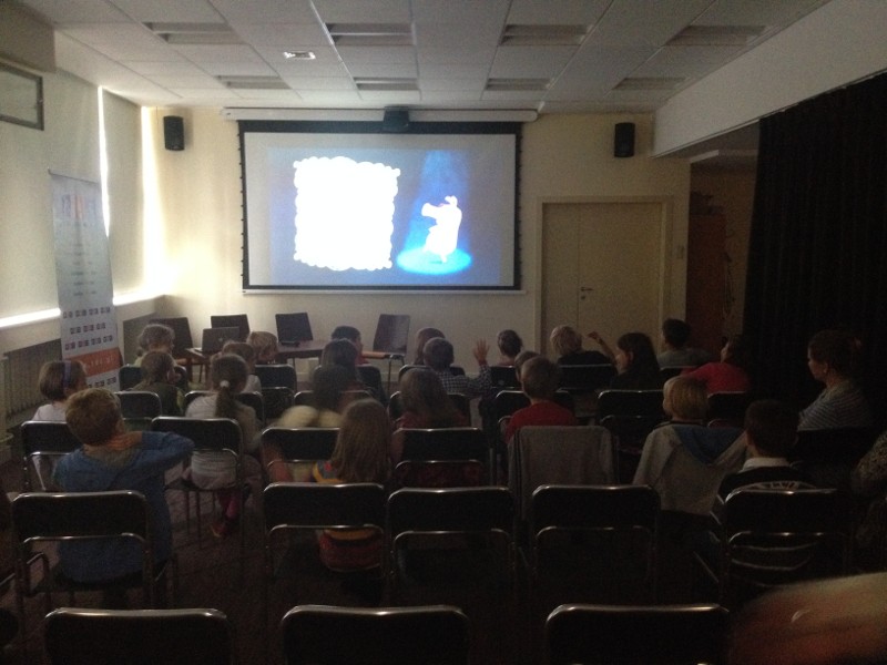 Grupa dzieci siedzi w rzędach krzeseł i ogląda animowany film wyświetlany na ekranie w słabo oświetlonym pomieszczeniu.