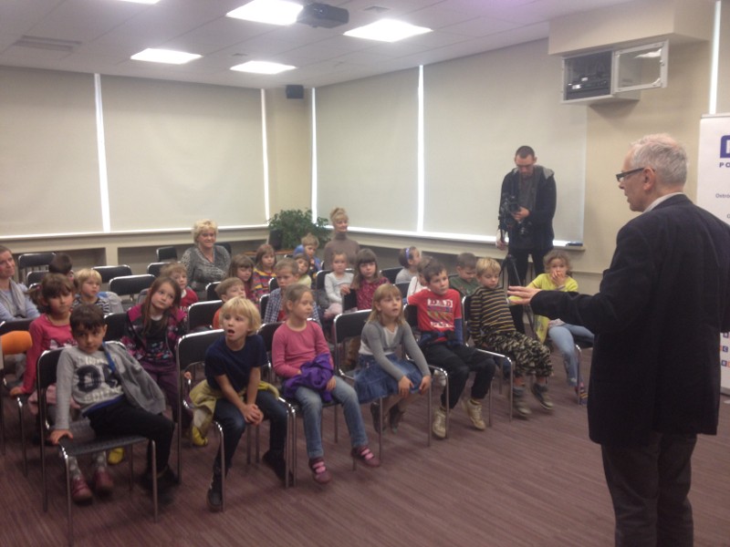 Mężczyzna stoi z przodu sali i przemawia do grupy małych dzieci siedzących na krzesłach, podczas gdy kilku dorosłych obserwuje z tyłu. Sceneria wydaje się być klasą lub salą konferencyjną z dużymi oknami.